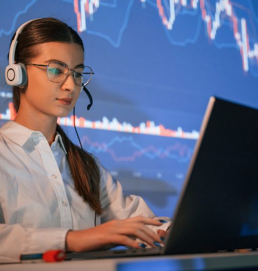 Sitting by laptop. Female stock broker is working indoors. Big wide display behind her.